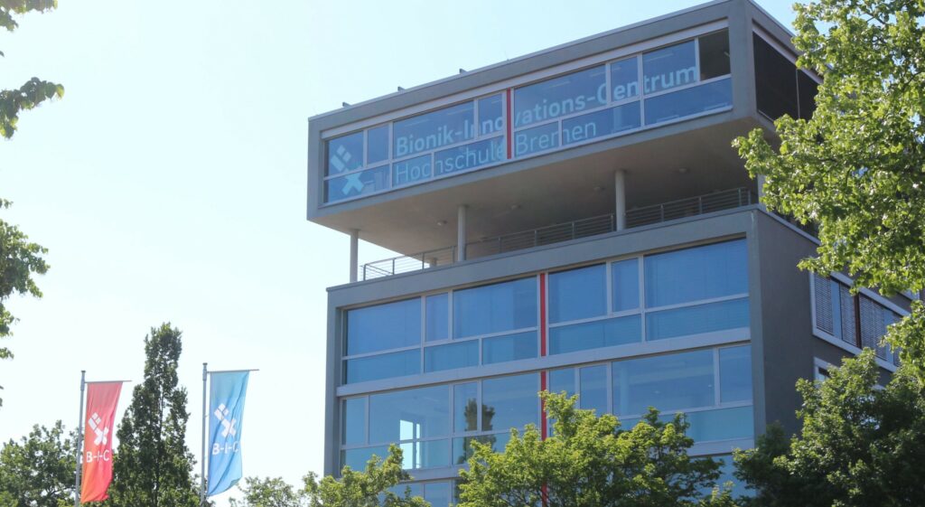 Außenansicht des Gebäudes des Bionik-Innovations-Centrums der Hochschule Bremen. Rechts und links, sowie unten am Rand stehen Bäume, zwei Flaggen links unten zeigen das B-I-C Logo. Das Gebäude ist grau und besteht aus Glas und Beton.
