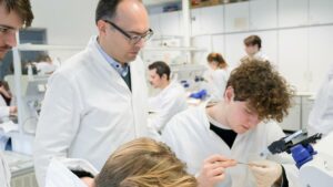 Eine helle Laborumgebung, im Vordergrund rechts sitzen zwei Studierende in weißen Laborkitteln. Sie präparieren etwas mit einer Pinzette. Vor ihnen steht ein Mikroskop. Ein Professor steht hinter ihnen und schaut ihnen über die Schultern. Im Hintergrund sind weitere Studierende, die ebenfalls im Labor beschäftigt sind.