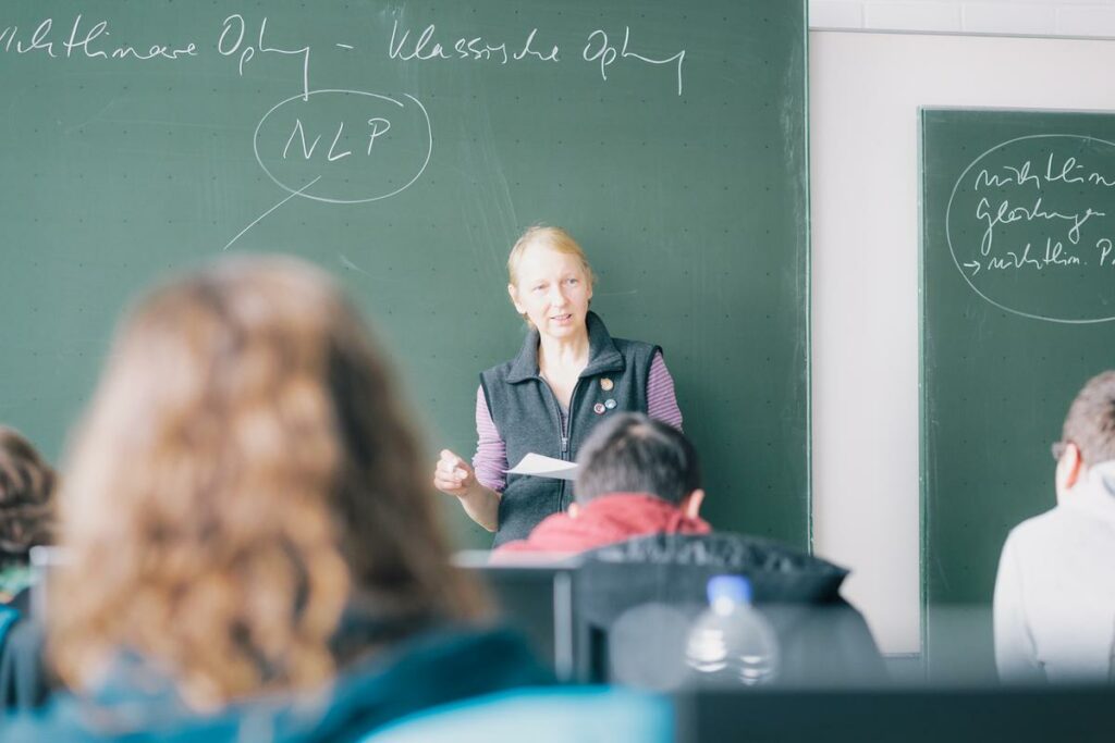 Eine Vorlesungssituation mit einer Professorin, die den Zuhörenden zugewandt mit dem Rücken zu einer grünen Tafel steht. Sie hält in einer Art Papier und in der anderen Hand ein Stück Kreide. Im Vordergrund verschwommen sieht man die Rücken der Zuhörenden.