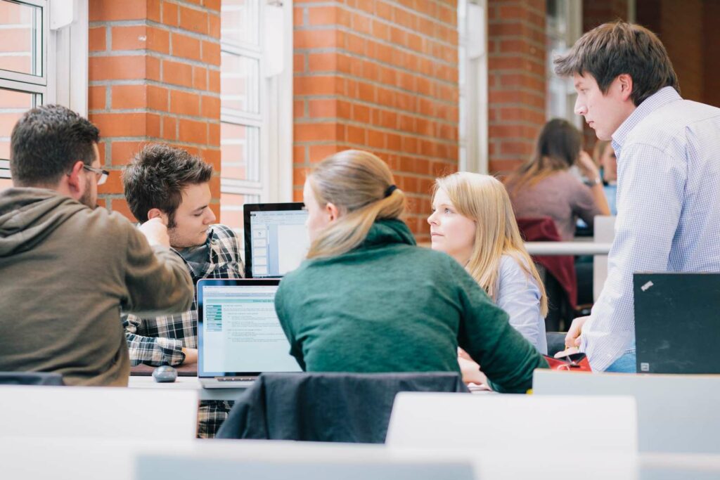 Eine Gruppe Studierender sitzt einander zugewandt um einen Tisch. Zwei Laptops stehen in ihrer Mitte und eine Person erklärt den anderen etwas und zeigt auf einen Laptopbildschirm.