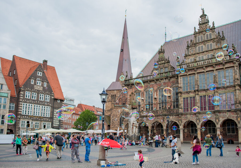 Buntes Treiben auf dem Bremer Marktplatz mit Blick auf das historische Rathaus im Hintergrund. Im Vordergrund schweben Seifenblasen durchs Bild.