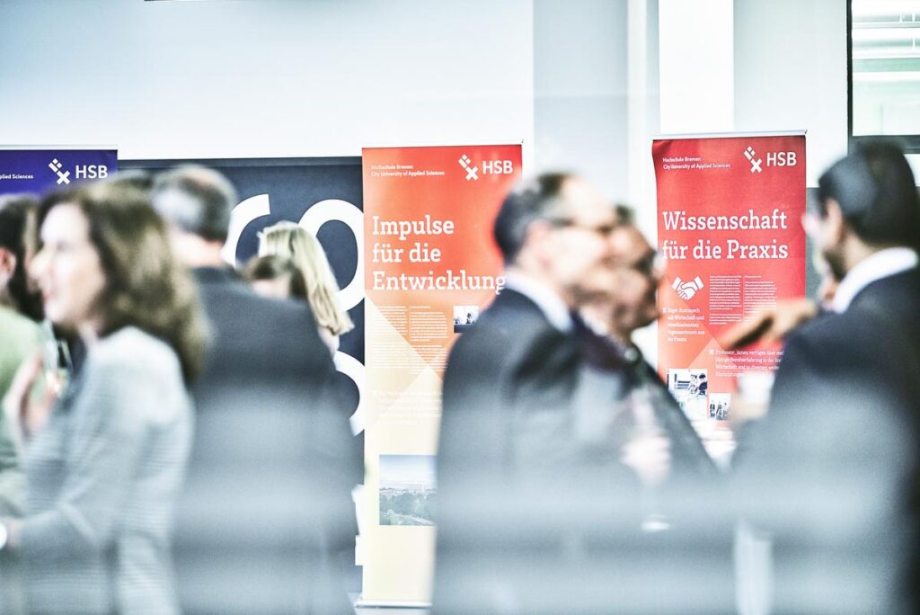 Im Vordergrund eine verschwommende Menschengruppe in Gesprächen. Im Hintergrund bunte Roll-ups der Hochschule Bremen.