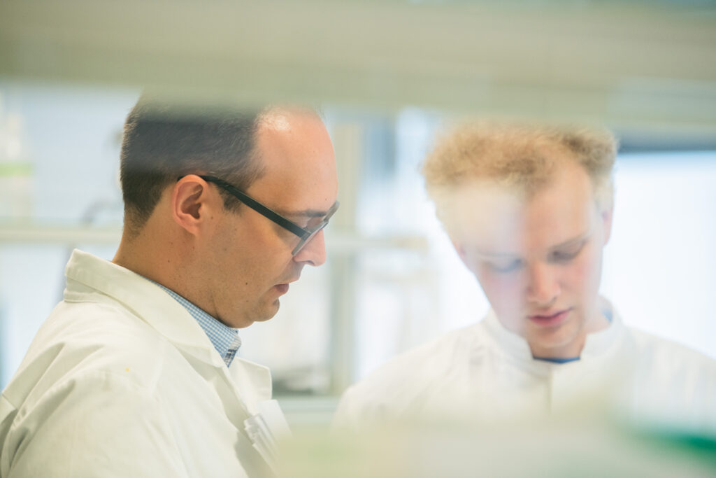 Seitliches Portrai einer Person auf der linken Bildseite. Die Person trägt einen Laborkittel, hat kurze dunkle Haare und trägt eine dunkle Brille. Eine zweite Person steht auf der rechten Bildseite. Diese Person trägt ebenfalls einen Laborkittel und hat helles kurzes Haar und schaut nach unten.