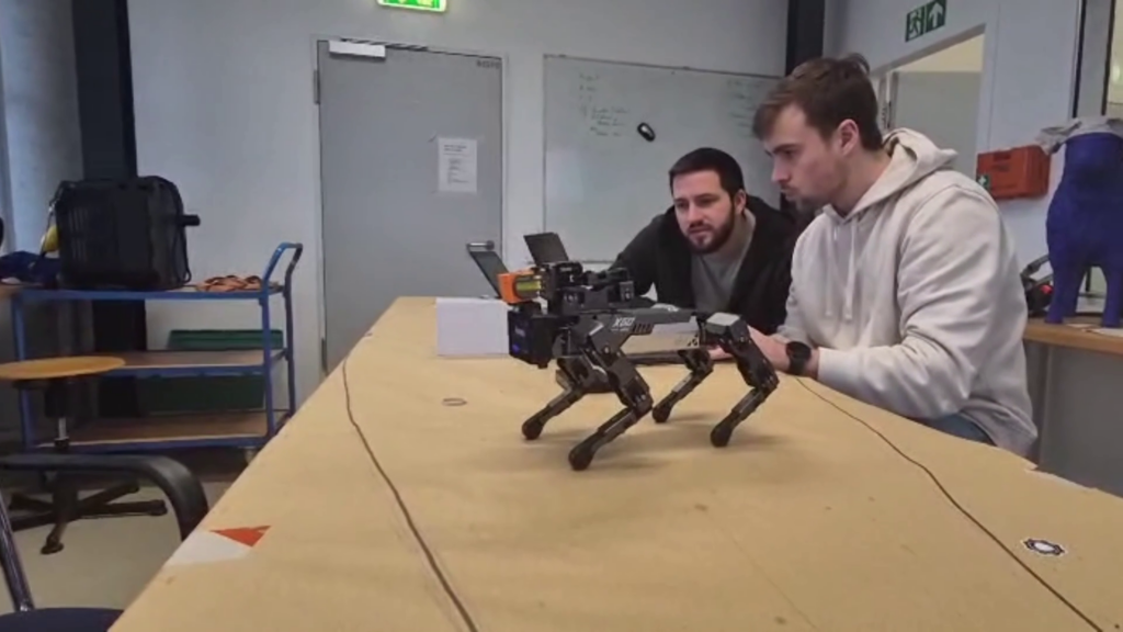 im Vodergrund ein grauer roboterhund auf einem tisch. im hintergrund dahinter zwei studierende vor einem laptop sitzend.