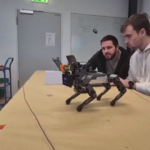 im Vodergrund ein grauer roboterhund auf einem tisch. im hintergrund dahinter zwei studierende vor einem laptop sitzend.