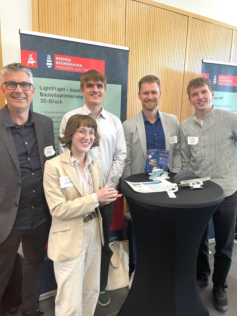 Eine Gruppe Menschen steht um einen Stehtisch und lacht stolz in die Kamera. Eine junge Frau präsentiert den Gewonnen Bre3DAward Pokal. Im Hintergrund steht das Roll-up des Projektteams.